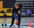 Person playing bocce indoors, focusing intensely while rolling a red ball on a gym floor.