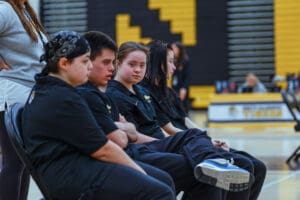 A group of young athletes sitting on chairs in a gymnasium, wearing team uniforms.