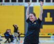 Woman in a black shirt smiling with arms raised in a gymnasium.