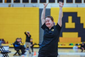 Woman in a black shirt smiling with arms raised in a gymnasium.