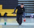 Person playing bocce indoors, wearing a black sports outfit, focused on rolling a red ball.