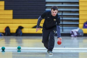 Person playing bocce indoors, wearing a black sports outfit, focused on rolling a red ball.