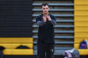 A person in a black sports outfit stands clapping in front of bleachers, focused and engaged in an indoor setting.