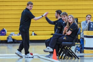 Team members in a gymnasium high-five while others watch, seated near a scoreboard, showing teamwork and engagement.
