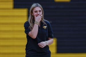 Young woman in black polo, hand on mouth, standing on yellow and black steps indoors.
