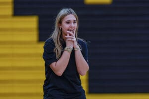 Young woman in black shirt with bracelets, standing in gym, hands clasped, expressing excitement or anticipation.