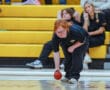 A focused bowler rolls a red ball on a court, with spectators watching from the bleachers in the background.