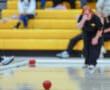Person preparing to release a red bocce ball on a gym floor with yellow bleachers in the background.