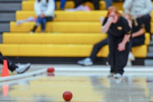 Person preparing to release a red bocce ball on a gym floor with yellow bleachers in the background.