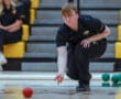 Person in black uniform rolling a red ball on an indoor court, focused and kneeling, with yellow seats in the background.