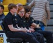 A group of young people in black uniforms sitting on a bench at an indoor sports event.