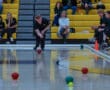 Person playing boccia in a gymnasium, with spectators seated on yellow bleachers in the background.