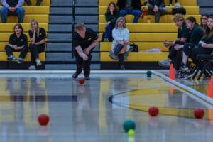Person playing boccia in a gymnasium, with spectators seated on yellow bleachers in the background.