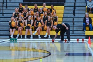 Person preparing to roll a red bocce ball on gym floor as cheerleaders watch, seated on yellow bleachers.