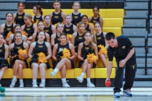A young man prepares to throw a red ball in a gym while a cheer team watches from the bleachers.