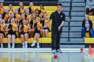 Teen participates in indoor sports activity with cheerleaders and audience watching in gymnasium.