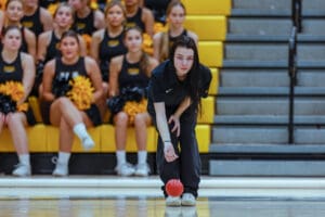 A person bowling with intense focus, surrounded by a cheering squad in a gymnasium.