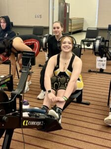 Smiling woman using a rowing machine indoors, surrounded by others exercising.