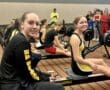 Participants at an indoor rowing competition, seated on ergometers, with spectators in the background.