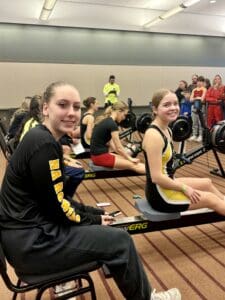 Participants at an indoor rowing competition, seated on ergometers, with spectators in the background.