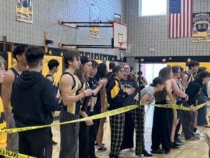 School athletes and students in a gym cheer for teammates during a sports event, with caution tape marking boundaries.