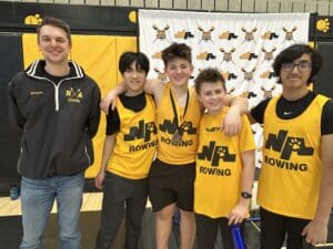 Youth rowing team poses with coach in yellow uniforms at a sports event backdrop.