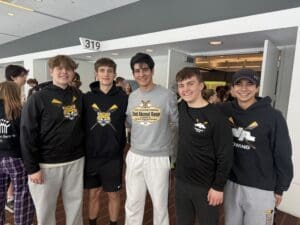 Group of five smiling rowers in team apparel at an alumni race event indoors.