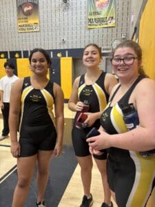 Three student athletes in black and yellow sports attire smiling in a gym setting.