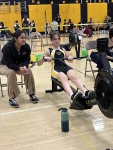 Person rowing on a machine with a coach beside them, focused on fitness training indoors.