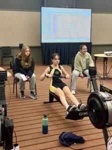 Teen girl rowing on an indoor machine during a competition, with results displayed on the screen behind her.