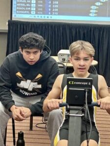 Two young men focusing on rowing competition, one using a Concept2 rowing machine, screen displaying results.
