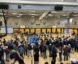 Crowded gym with athletes and spectators at an indoor sports event, featuring rowing machines and scoreboard.