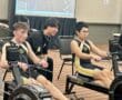 Two athletes in team uniforms rowing on indoor machines during a competition.