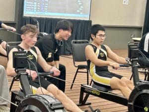 Two athletes in team uniforms rowing on indoor machines during a competition.