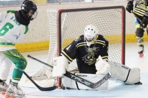 Youth hockey goalie defends the goal against an approaching player, wearing green and white, during a match on the ice.