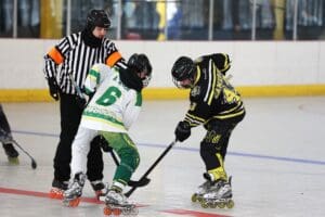 Two inline hockey players face off as the referee prepares for the puck drop on the rink, wearing safety gear.
