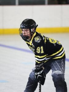 Hockey player in black and yellow jersey focuses during a game on ice rink, wearing a helmet and holding a stick.