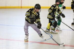 Youth roller hockey player in action, wearing a black and yellow jersey, on an indoor rink.
