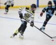 Youth roller hockey game, player in white jersey skating and controlling puck indoors.