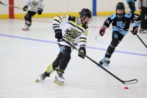 Youth roller hockey game, player in white jersey skating and controlling puck indoors.