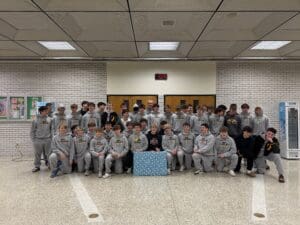 A large group of young athletes in matching track suits pose inside a building with a blue wrapped gift in front.