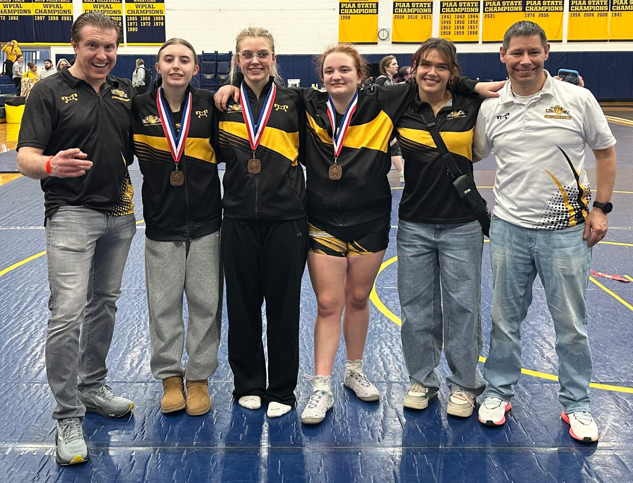 Group of athletes and coaches celebrating with medals in a gymnasium with championship banners in the background.