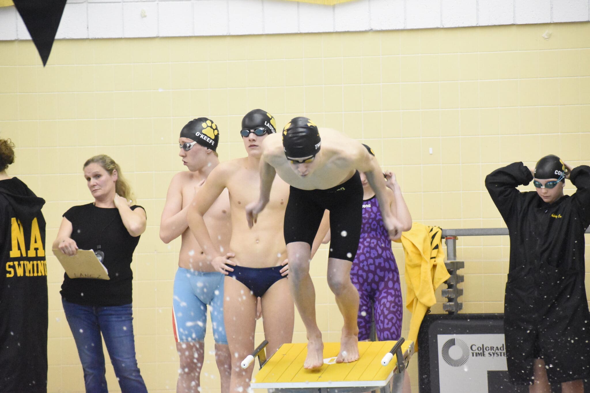 Swimmer dives off starting block at indoor pool competition; teammates and coach watch nearby.