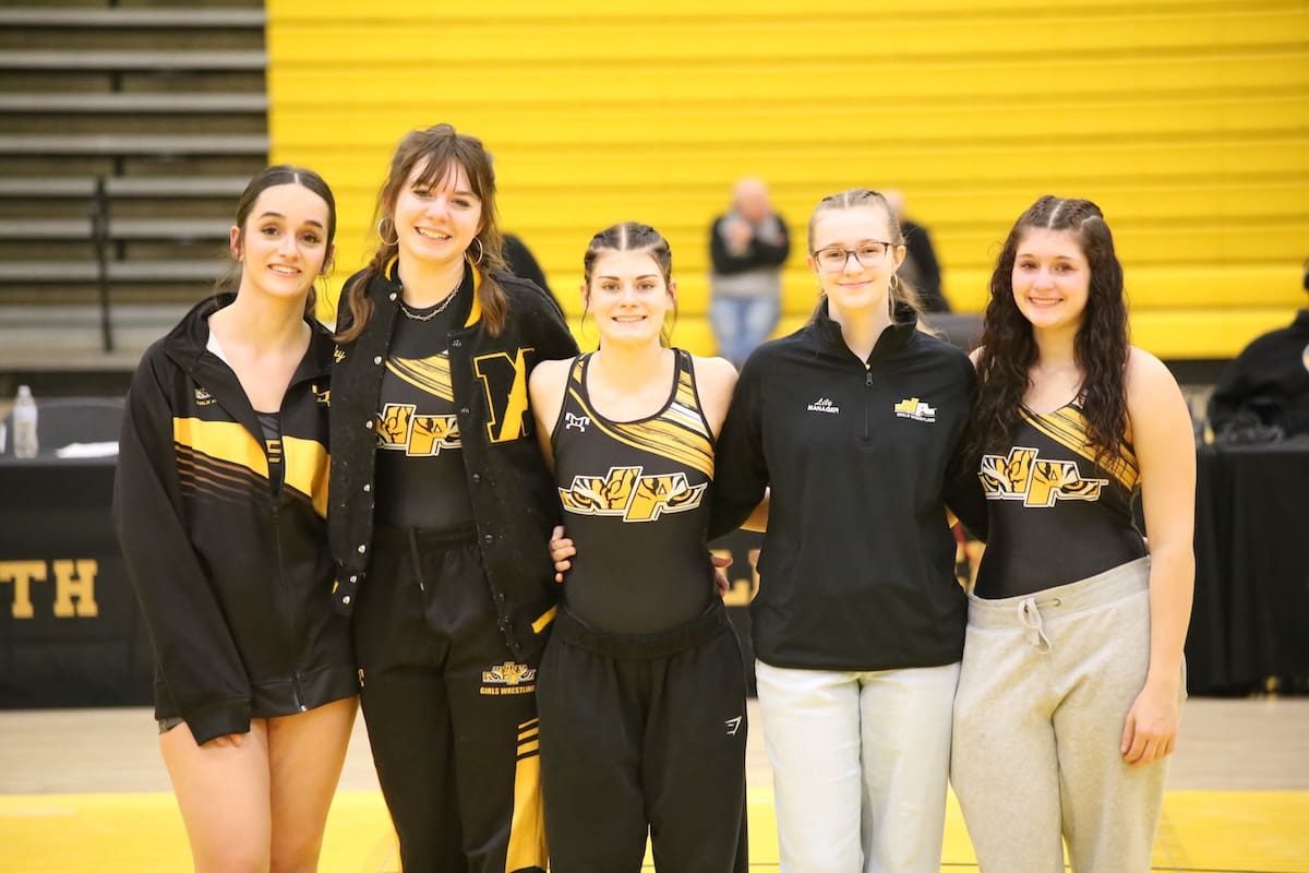 Five female wrestlers in team uniforms posing together indoors.