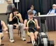 Teen rowing competition indoors with participants focused on the machine and judges observing in the background.