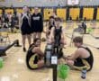 Indoor rowing team training together in a school gym, wearing matching uniforms and using rowing machines.