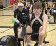 Teen rowing athlete competing on Concept2 machine in a gym, surrounded by coach and teammates.