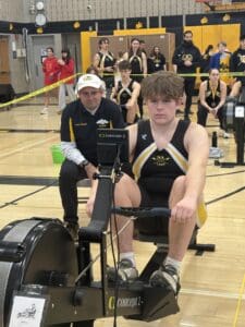 Teen rowing athlete competing on Concept2 machine in a gym, surrounded by coach and teammates.