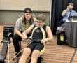 Young athlete rowing on an indoor machine with a coach cheering on in a competitive event setting.