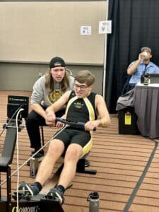 Young athlete rowing on an indoor machine with a coach cheering on in a competitive event setting.